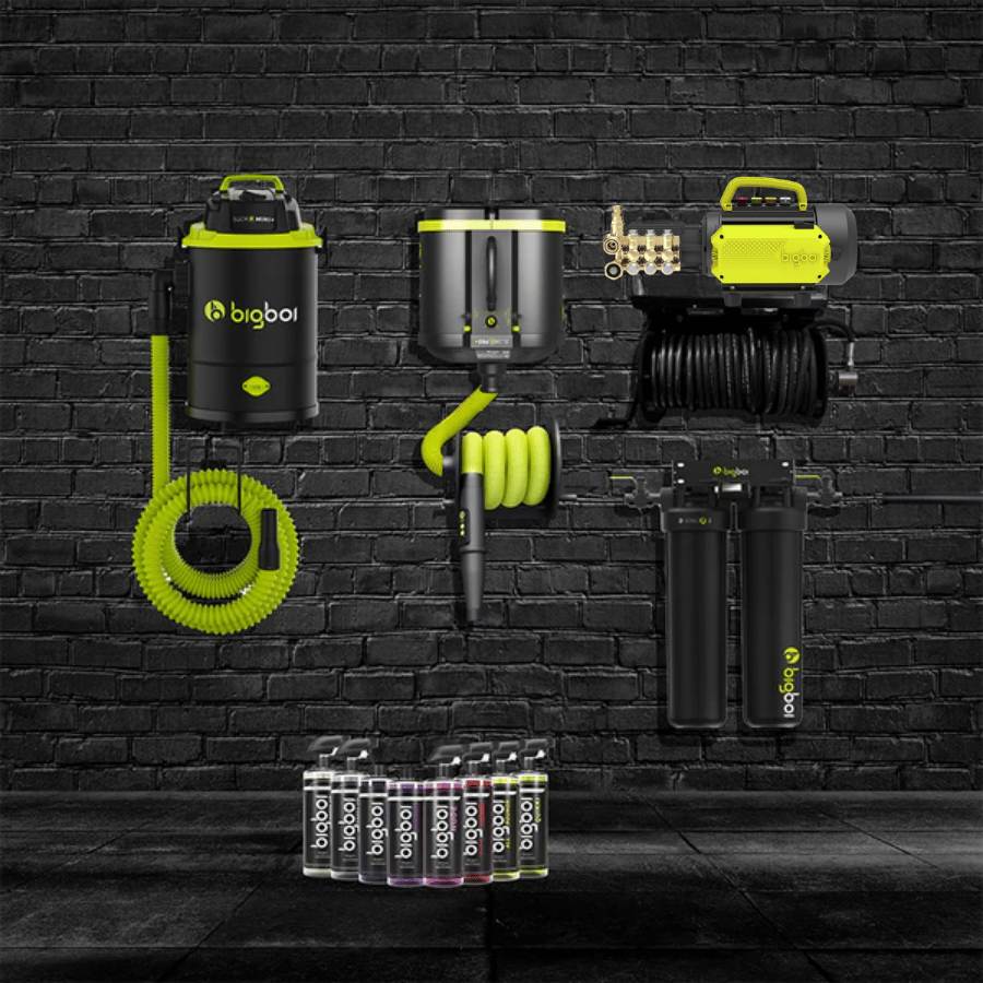 Collection of BigBoi branded equipment including a pressure washer, hose, and chemicals against a black brick wall.